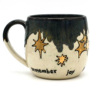 May include: A ceramic mug with a cream-colored base and a dark blue-green top. Gold star designs are scattered across the mug, with the words "remember joy" in black script. The handle is black.