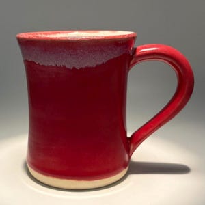 May include: A ceramic mug with a glossy red glaze that transitions to a lighter, textured white rim. The mug features a curved handle on the right side and a cylindrical body that tapers slightly toward the base, which is left in a natural cream color.