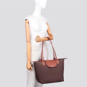 May include: A dark brown nylon tote bag featuring two long shoulder straps and a fold-over flap with a snap button closure. The bag has a rectangular silhouette with a smooth, solid-coloured exterior and contrasting tan leather-like trim.