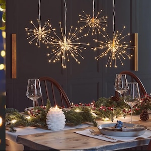 May include: A decorated dining table with starburst lights suspended from above. The table is set with wine glasses, a white candle, and a garland of greenery and berries. A plate and cutlery are also visible.