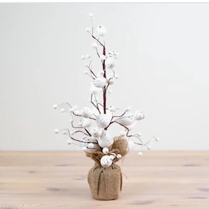 May include: An artificial tree featuring white speckled egg-shaped ornaments and small white berries on brown branches. The base is wrapped in burlap. The tree is set on a light-coloured wooden surface, creating a rustic look.