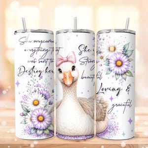 May include: A white tumbler featuring a goose with a pink bow, adorned with purple and white floral designs. The tumbler displays the text "She overcame everything that was sent to destroy her" and "She is strong, beautiful, loving & graceful."