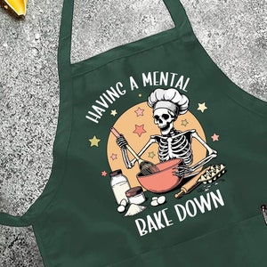 May include: Dark green apron with the text "HAVING A MENTAL BAKE DOWN" and a skeleton chef graphic. The apron has a pocket and adjustable neck strap. The image also includes pasta, tomatoes, olives, and other cooking ingredients.