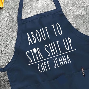 May include: Navy blue apron with white text that reads "About to Stir Shit Up" and "Chef Jenna".