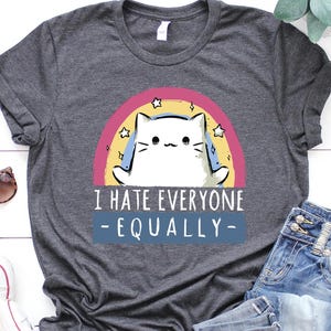 May include: Dark gray t-shirt with a cat graphic inside a rainbow and the text "I HATE EVERYONE - EQUALLY-". The shirt is paired with blue jeans, white sneakers, and sunglasses.