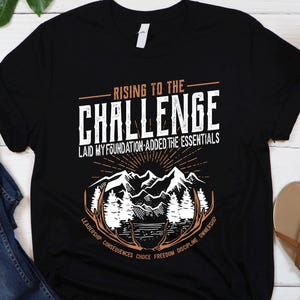 May include: Black t-shirt with a white graphic design that says "Rising to the Challenge Laid My Foundation - Added the Essentials" with a mountain range and antlers. The text below the graphic says "Leadership Confidence Choice Freedom Drive Me Everytime".