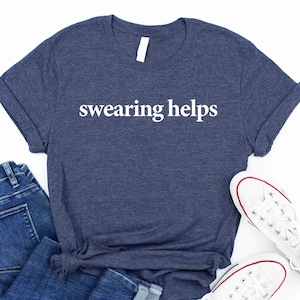 May include: Navy blue t-shirt with the text "swearing helps" in white, paired with blue jeans and white sneakers. The t-shirt is folded, and the sneakers have red accents.
