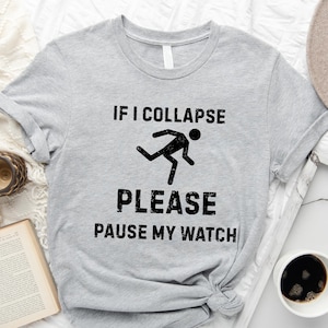 May include: Gray t-shirt with black text that reads "IF I COLLAPSE PLEASE PAUSE MY WATCH". A stick figure is running in the text. The shirt is on a white surface with a book, a clock, a cup of coffee, and a hat.