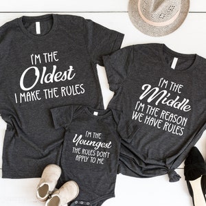 May include: Three dark gray t-shirts and a onesie with white text that reads "I'M THE Oldest I MAKE THE RULES", "I'M THE Middle I'M THE REASON WE HAVE RULES", and "I'M THE Youngest THE RULES DON'T APPLY TO ME". Accessories include sunglasses, shoes, and a hat.