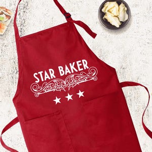 May include: A red apron with the text "STAR BAKER" in white script, adorned with decorative flourishes and three stars. The apron has two pockets and a neck strap. Sushi, limes, and ginger are arranged around the apron.