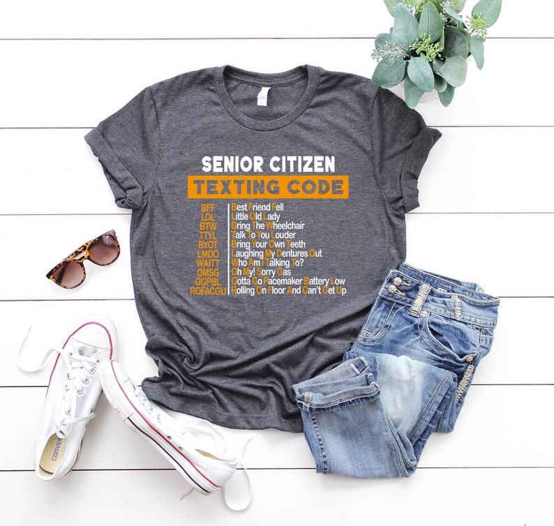 Pu&ograve; includere: Una maglietta grigia con il testo "Senior Citizen Texting Code" in arancione. Il testo &egrave; un elenco di abbreviazioni e i loro significati, come BFF, LOL, BTW, TYL, BYOT, LMDO, WAITT, OMSG, GGPBL e ROFACGU.