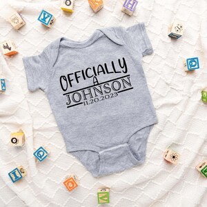May include: Gray baby onesie with the text "Officially a Johnson 11.20.2023" printed in black.