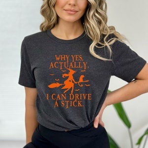 Może przedstawiać: Szary t-shirt z pomarańczowym nadrukiem przedstawiającym czarownicę na miotle z czarnym kotem. Tekst na koszulce brzmi "Why yes, actually. I can drive a stick."