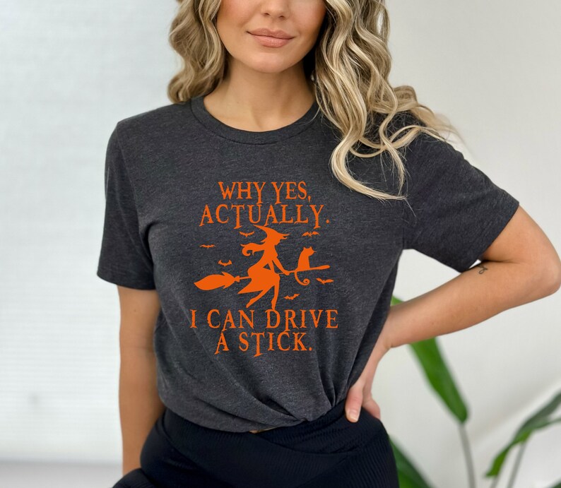 Może przedstawiać: Szary t-shirt z pomarańczowym nadrukiem przedstawiającym czarownicę na miotle z czarnym kotem. Tekst na koszulce brzmi "Why yes, actually. I can drive a stick."