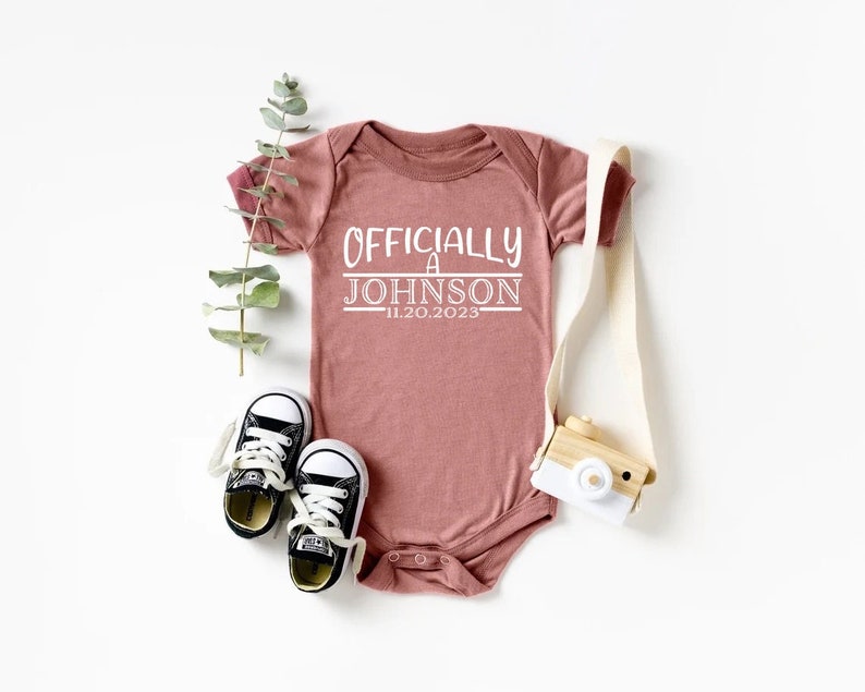 May include: A pink baby onesie with the text "Officially a Johnson - 11.20.2023" in white.