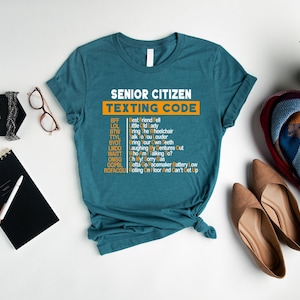 Pu&ograve; includere: Maglietta blu turchese con il testo "Senior Citizen Texting Code" in arancione e bianco. La maglietta elenca le comuni abbreviazioni di messaggi di testo degli anziani e i loro significati.