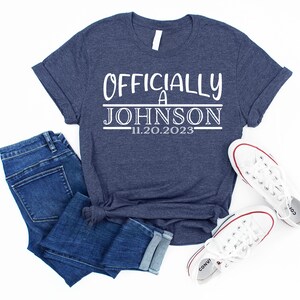 May include: A navy blue t-shirt with white text that reads "Officially a Johnson 11.20.2023". The shirt is laid flat on a white surface with a pair of white sneakers and blue jeans.