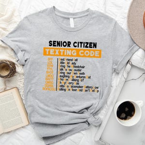 Pu&ograve; includere: Una maglietta grigia con il testo "Senior Citizen Texting Code" in nero e un elenco di abbreviazioni e i loro significati in arancione. Le abbreviazioni includono BFF, LOL, BTW, TTYL, BYOT, UMDO, WAITE, OMSG, GOPEL, ROFACCU.