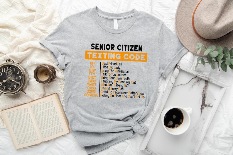 Pu&ograve; includere: Una maglietta grigia con il testo "Senior Citizen Texting Code" in nero e un elenco di abbreviazioni e i loro significati in arancione. Le abbreviazioni includono BFF, LOL, BTW, TTYL, BYOT, UMDO, WAITE, OMSG, GOPEL, ROFACCU.