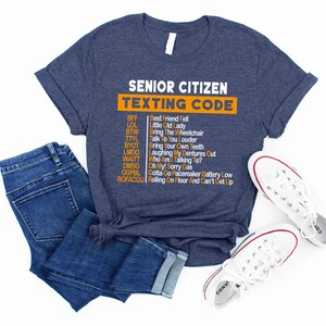 Pu&ograve; includere: Una maglietta blu navy con una grafica bianca che dice "Senior Citizen Texting Code" e elenca le comuni abbreviazioni di messaggi di testo degli anziani e i loro significati in testo arancione.