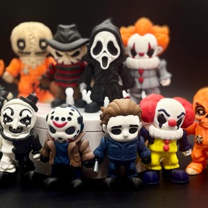 May include: A group of small, colourful figurines representing horror film characters. The figures are detailed, with recognisable features like masks and costumes. Characters include a Predator, a clown, and Michael Myers. The figurines are on display.