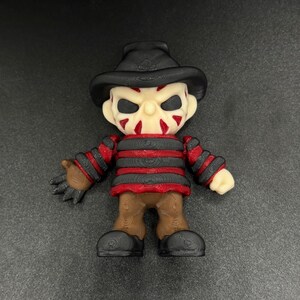 Pode incluir: Uma pequena figura listrada de vermelho e preto com um chap&eacute;u marrom e um rosto branco com marcas vermelhas. A figura tem uma express&atilde;o amea&ccedil;adora e est&aacute; segurando uma faca na m&atilde;o direita.
