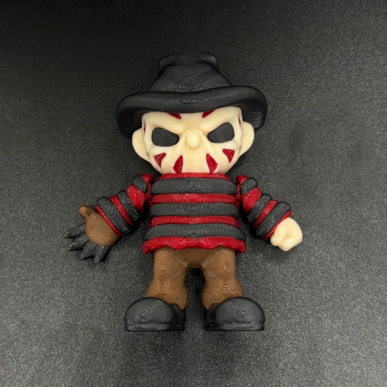 Pode incluir: Uma pequena figura listrada de vermelho e preto com um chap&eacute;u marrom e um rosto branco com marcas vermelhas. A figura tem uma express&atilde;o amea&ccedil;adora e est&aacute; segurando uma faca na m&atilde;o direita.