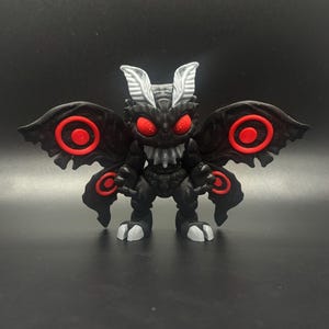 May include: A black figurine of a creature with large wings, red eyes, and a menacing expression. The wings have red circular accents. The figurine has white accents on the feet and head. The overall design is reminiscent of a moth.