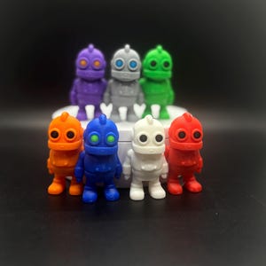 May include: A collection of colorful robot figurines in various colors, including purple, gray, green, orange, blue, white, and red. Each robot has a distinct design with round heads and body shapes. The robots are arranged on a white platform.