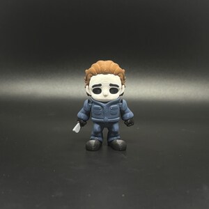 May include: A small figurine of Michael Myers from the Halloween horror film series. The figure is wearing a blue jumpsuit and a white mask with black eye holes. It is holding a small, white knife. The figure has brown hair.