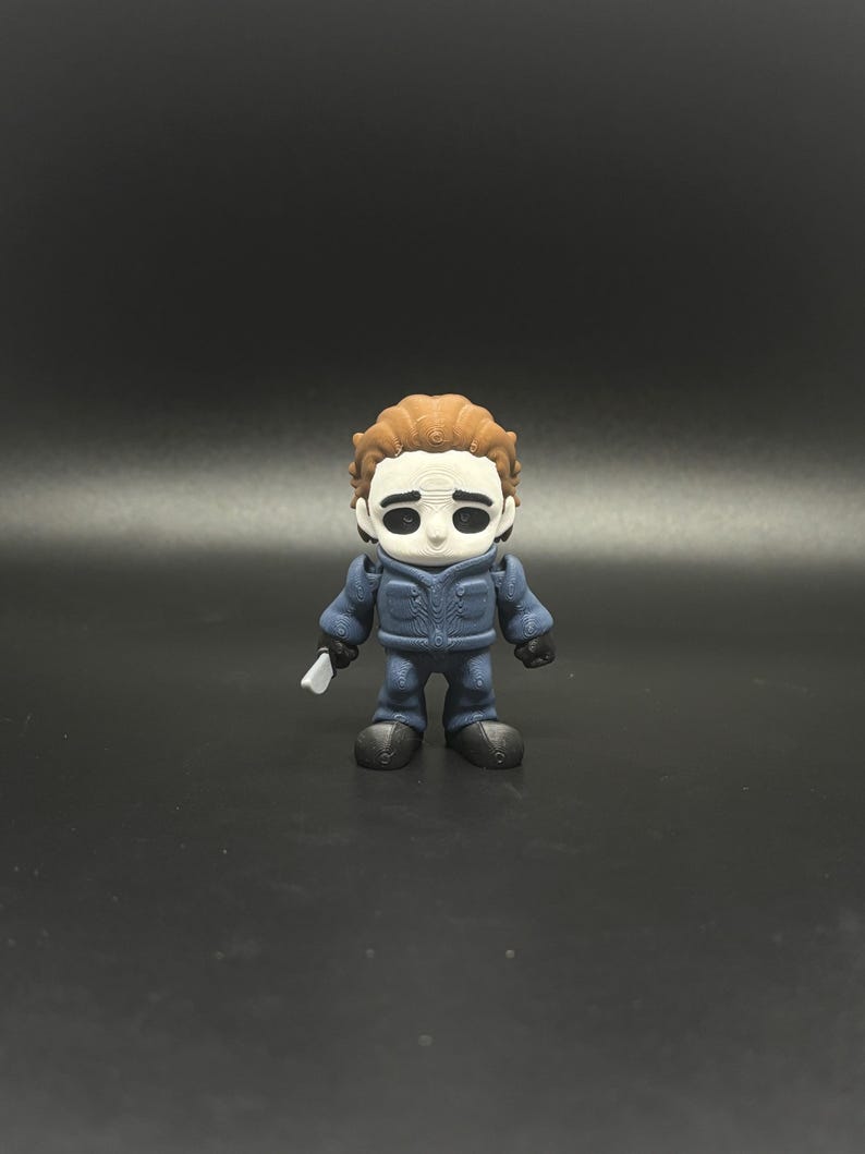 May include: A small figurine of Michael Myers from the Halloween horror film series. The figure is wearing a blue jumpsuit and a white mask with black eye holes. It is holding a small, white knife. The figure has brown hair.