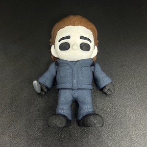 May include: A 3D-printed figurine of Michael Myers from the Halloween horror film series. The figure is wearing a blue jumpsuit and black gloves and shoes, and is holding a knife. The figure has a white mask and brown hair.
