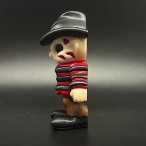 Pode incluir: Uma camisa listrada de vermelho e preto com um chap&eacute;u preto e cal&ccedil;as marrons. A figura tem um rosto branco com marcas vermelhas. Esta &eacute; uma figura impressa em 3D de um personagem de filme de terror.