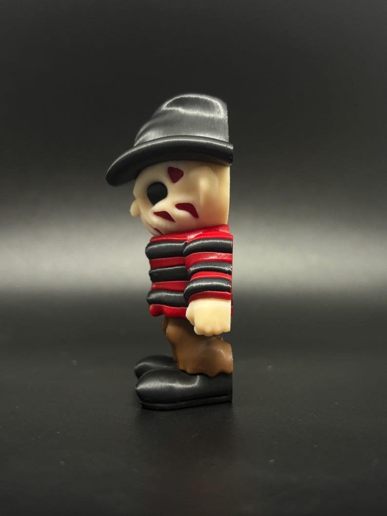 Pode incluir: Uma camisa listrada de vermelho e preto com um chap&eacute;u preto e cal&ccedil;as marrons. A figura tem um rosto branco com marcas vermelhas. Esta &eacute; uma figura impressa em 3D de um personagem de filme de terror.