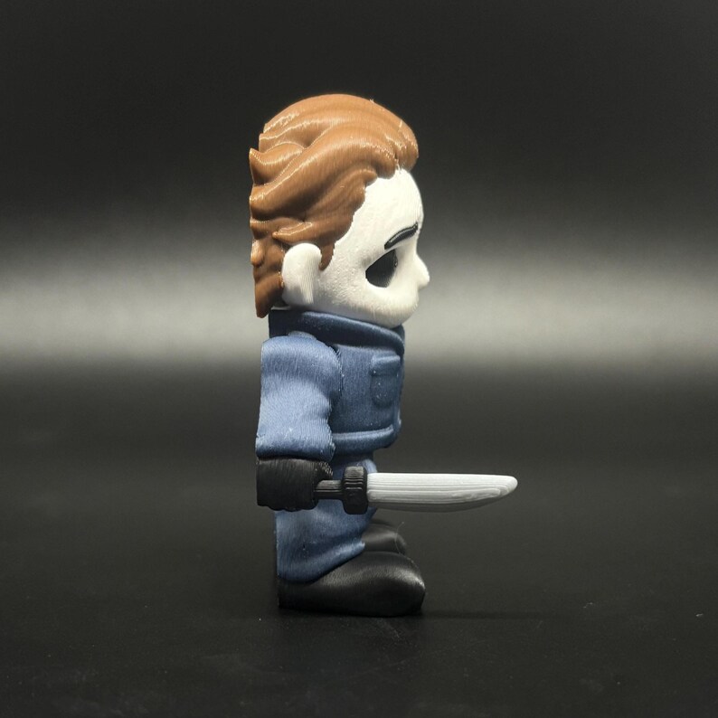 May include: A small, stylised figurine of a horror movie character. The figure has a white mask, brown hair, and wears a blue jumpsuit and black gloves. It holds a silver knife.