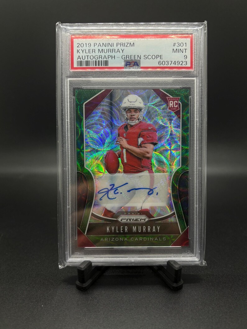 May include: A graded trading card of Kyler Murray, a football player for the Arizona Cardinals. The card is from the 2019 Panini Prizm Green Scope Autograph set and is graded a 9 by PSA. The card features a holographic design with a green background and a picture of Murray in his Cardinals uniform. The card also includes Murray's autograph.