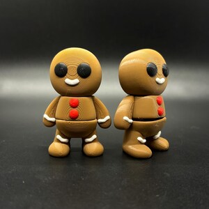 May include: Two brown gingerbread man figurines with black eyes, white smiles, and red heart-shaped buttons. The figurines have white icing details on their arms and legs, and are standing on a black surface.