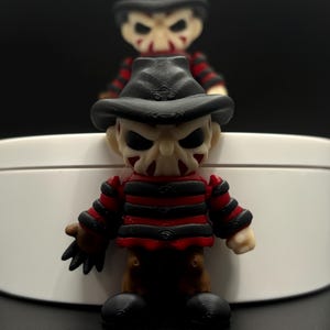 Pode incluir: Uma figura de Freddy Krueger com listras vermelhas e pretas, um chap&eacute;u preto e uma cara assustadora. A figura est&aacute; em p&eacute; sobre uma superf&iacute;cie branca.