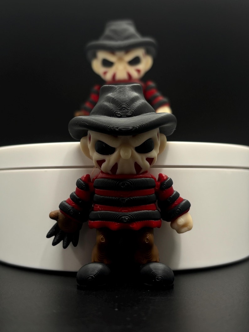 Pode incluir: Uma figura de Freddy Krueger com listras vermelhas e pretas, um chap&eacute;u preto e uma cara assustadora. A figura est&aacute; em p&eacute; sobre uma superf&iacute;cie branca.