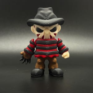 Pode incluir: Uma pequena figura listrada de vermelho e preto com um chap&eacute;u marrom e uma m&aacute;scara branca. A figura tem uma express&atilde;o amea&ccedil;adora e est&aacute; segurando uma faca na m&atilde;o direita.