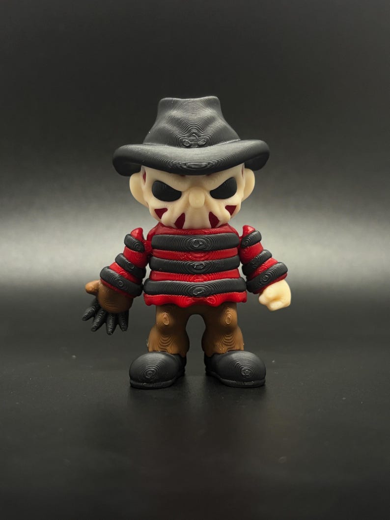 Pode incluir: Uma pequena figura listrada de vermelho e preto com um chap&eacute;u marrom e uma m&aacute;scara branca. A figura tem uma express&atilde;o amea&ccedil;adora e est&aacute; segurando uma faca na m&atilde;o direita.