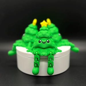 May include: A group of green Christmas tree figurines with smiling faces and yellow star toppers. The trees have articulated legs and are arranged on a white circular base. The background is dark.