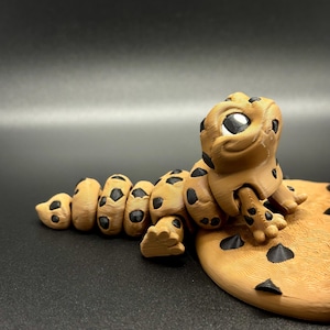 May include: A 3D printed figurine of a brown and black leopard gecko sitting on a brown and black cookie. The gecko has large, black eyes and a smiling expression.