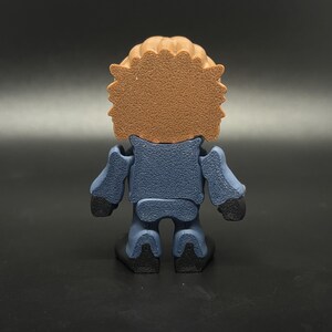 May include: A small, stylized figurine viewed from the back. The figure has a brown, textured head and a blue body with black hands and feet. The figure is standing on a black surface.