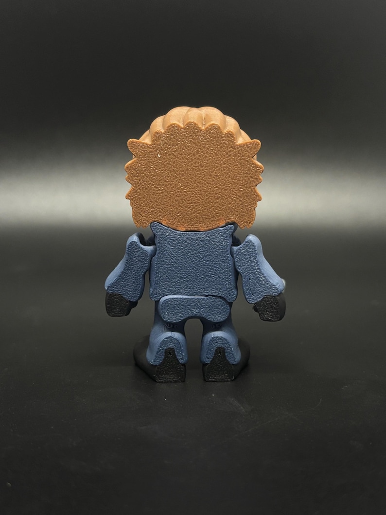 May include: A small, stylized figurine viewed from the back. The figure has a brown, textured head and a blue body with black hands and feet. The figure is standing on a black surface.