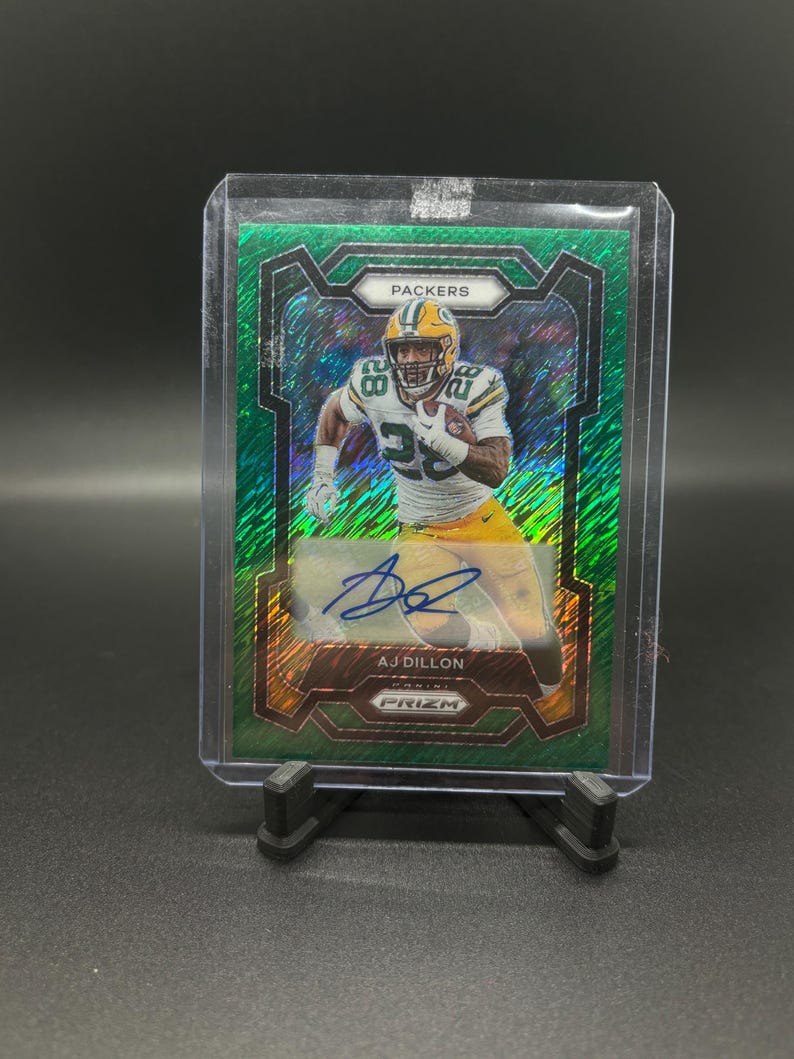 May include: Green, gold and silver football trading card featuring a player in a green and gold uniform with the text "PACKERS" and "AJ DILLON" on the card. The card is a Prizm card.