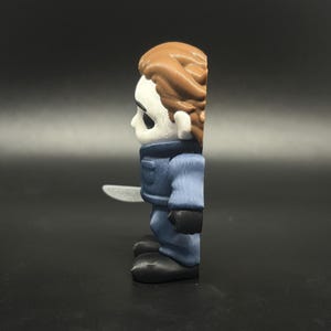 May include: A side profile of a small, stylised figure of a horror movie character. The figure has a white mask, brown hair, and wears a blue jumpsuit with black gloves and shoes. It holds a silver knife.