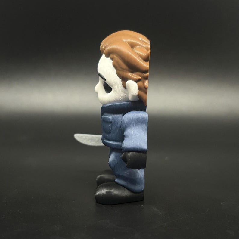 May include: A side profile of a small, stylised figure of a horror movie character. The figure has a white mask, brown hair, and wears a blue jumpsuit with black gloves and shoes. It holds a silver knife.