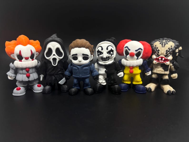 May include: A group of small figurines depicting characters from horror films. The figures include a clown with orange hair, a figure in a black robe with a white mask, and a figure in a blue jumpsuit. They are on a black surface.