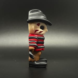 Pode incluir: Uma figura de Freddy Krueger com listras vermelhas e pretas, um chap&eacute;u preto e um rosto castanho. A figura &eacute; feita de pl&aacute;stico e tem cerca de 7,5 cm de altura.
